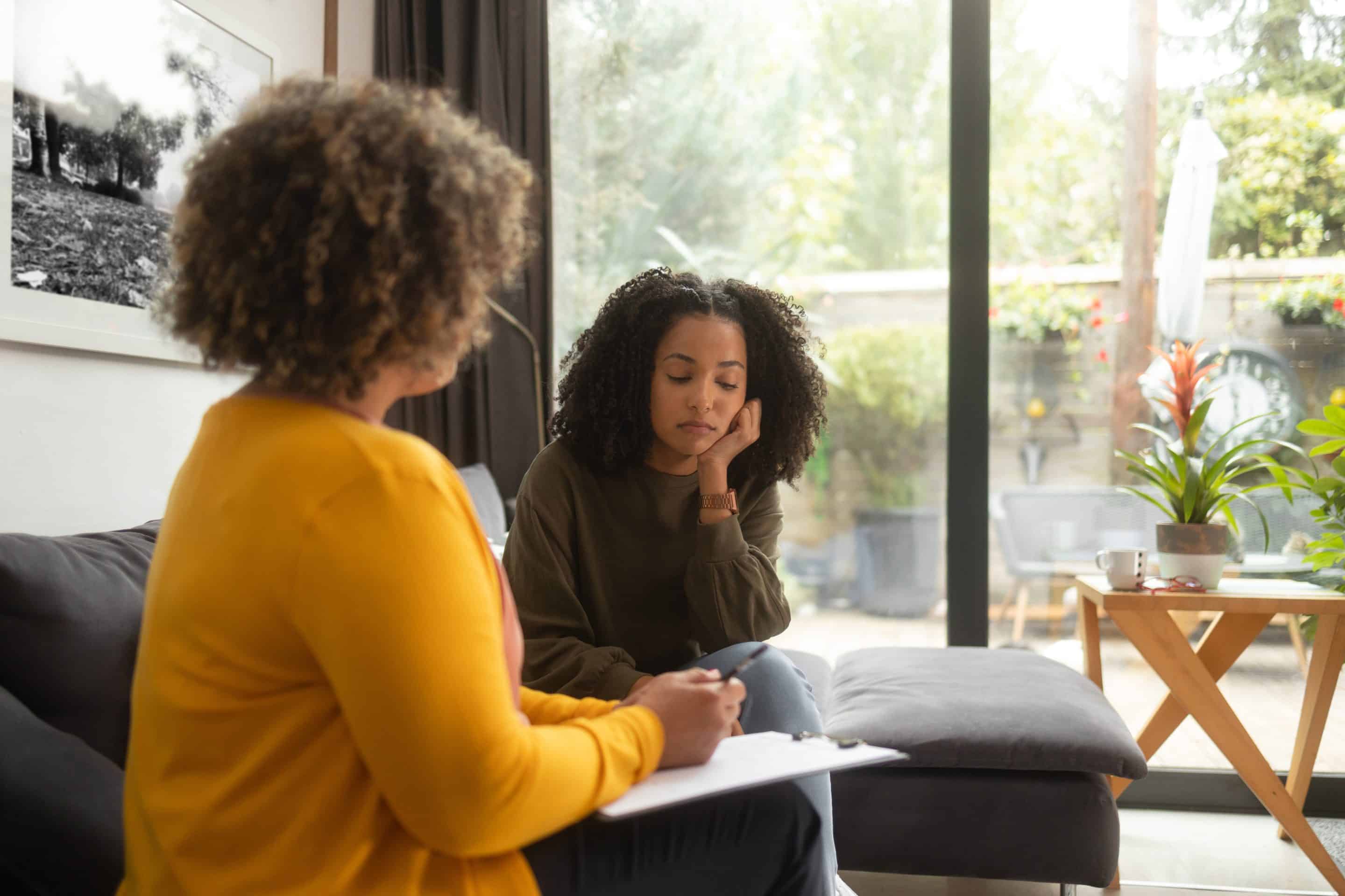 An African-American teen girl sits on a couch and talks to the unrecognizable female psychotherapist at her office. The girl rests her head on the hand and looks like she’s uncomfortable.